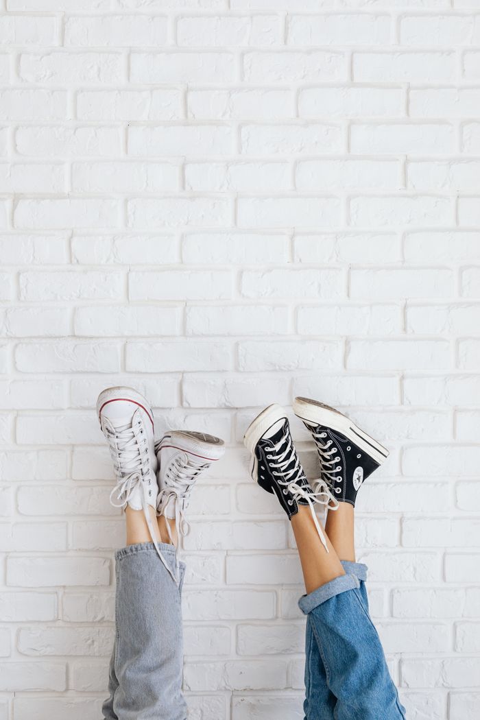 our-story Two pairs of stylish sneakers held up against a white brick wall, showcasing casual fashion.